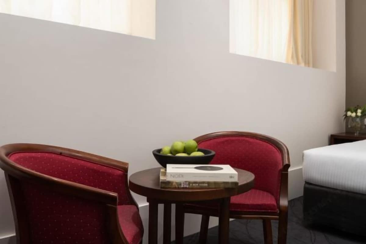 Habitación de hotel con cama, una mesa redonda de madera con un cuenco de fruta y un libro, y dos sillones tapizados en rojo.