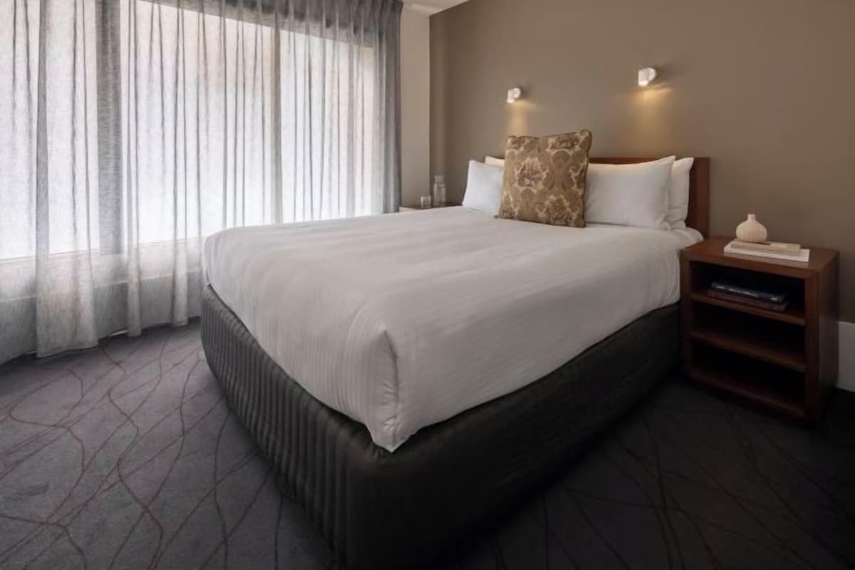 Una habitación de hotel con una cama grande, una mesita de noche, una lámpara y cortinas translúcidas.