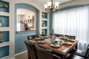 Een eetkamer met een grote tafel voor zes personen, omringd door stoelen. Daarboven hangt een kroonluchter en aan de rechterkant bevindt zich een raam met dunne gordijnen.