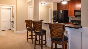 A kitchen with wooden cabinets, a black refrigerator, and a microwave. There are two wooden chairs with black cushions.