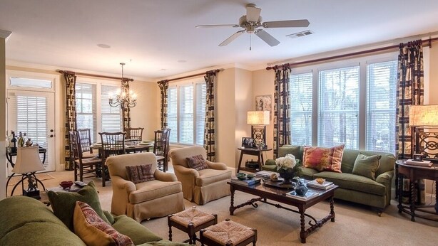 A spacious living room with a high ceiling fan, multiple seating options including a sofa, chairs, and a coffee table. Large windows allow natural light to fill the room.