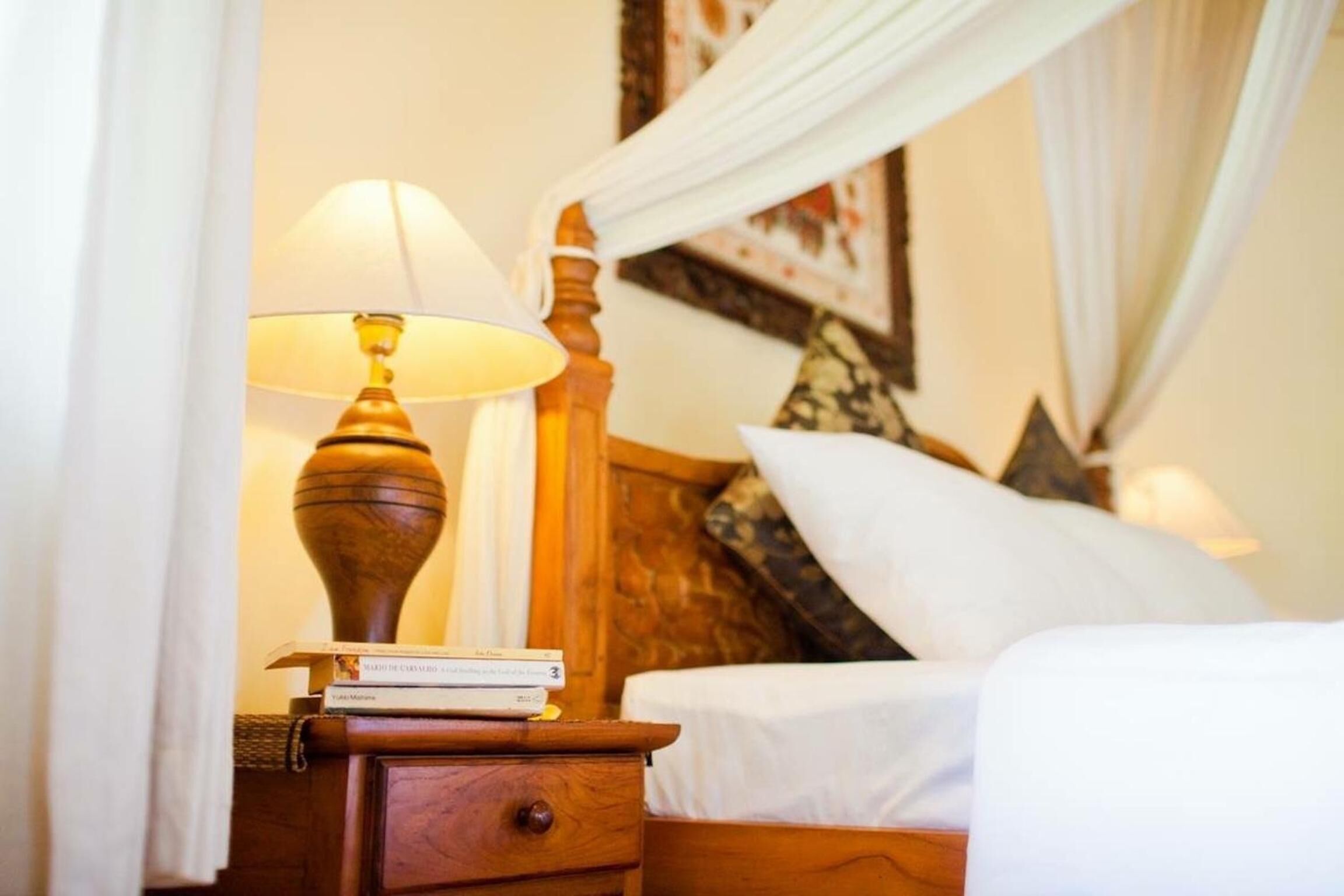 A neatly made bed with white linens, a wooden nightstand with a lamp, and a bedside table with books.