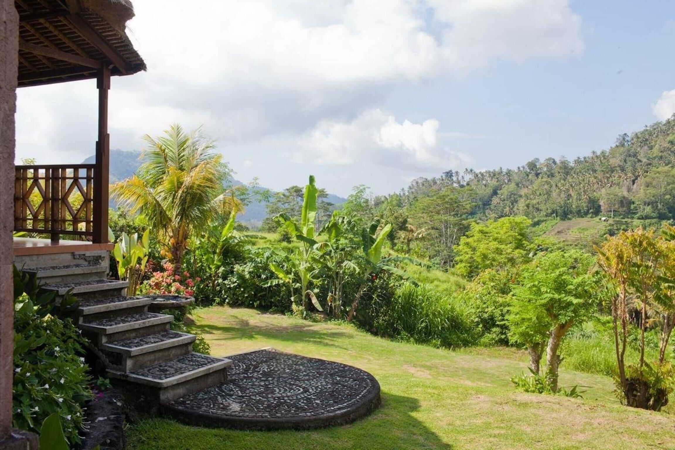 Un jardín con una escalera de piedra que lleva a una estructura de madera, rodeado de abundante vegetación y vistas a un paisaje montañoso.