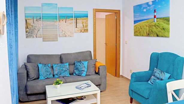 A living room with a grey sofa, a blue armchair, and a coffee table. There are four wall-mounted paintings depicting beach scenes and a lighthouse. - Apartment close to the beach (TARIFA)
