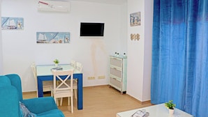 A living room with a blue sofa, a dining table with chairs, a small white table with a plant, and a TV mounted on the wall. - Apartment close to the beach (TARIFA)