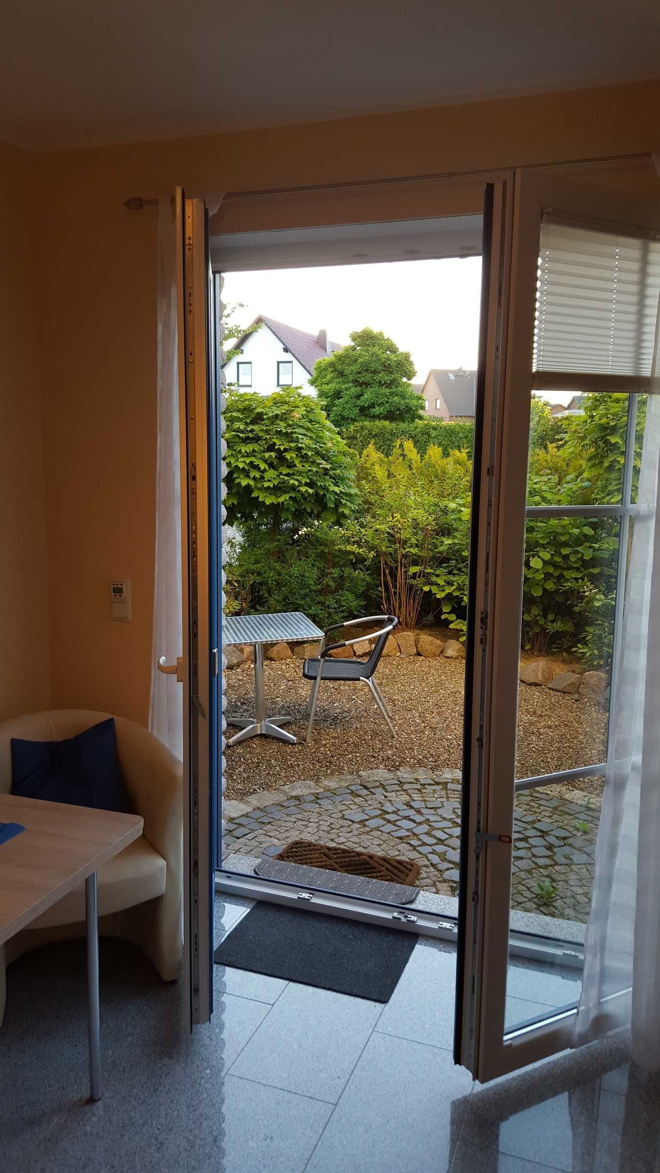 Ein Zimmer mit einer Schiebetür, die auf eine Terrasse mit Tisch und Stuhl, eine Fußmatte und einen Blick auf ein Haus und Grünflächen führt.