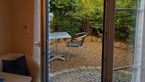 A room with a sliding glass door leading to a patio with a table and chair, a doormat, and a view of a house and greenery.