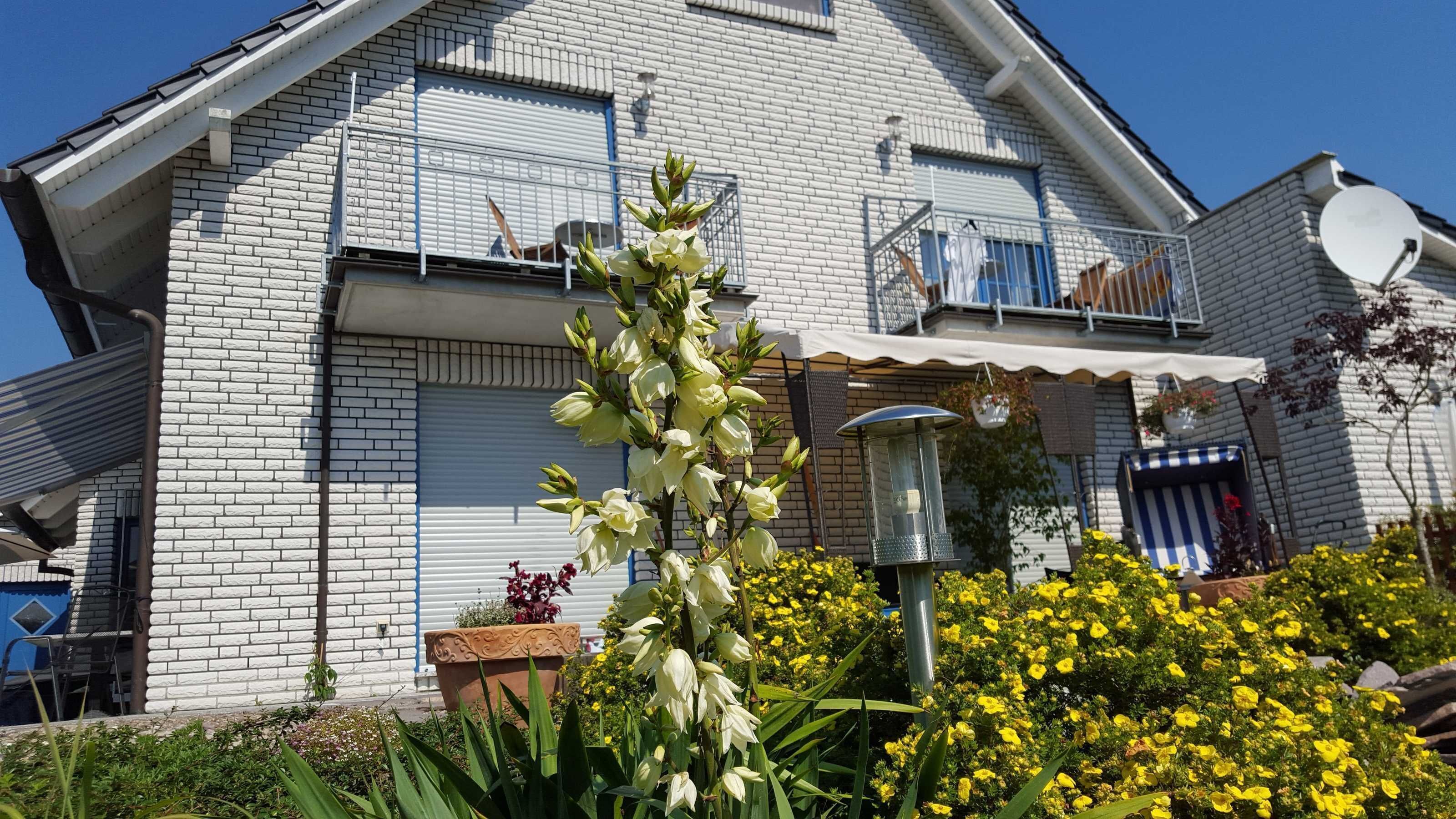 Ein weißes Backsteinhäuschen mit Satellitenschüssel, einem Balkon mit Geländer und einem Garten mit gelben Blumen.