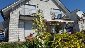 A white brick house with a satellite dish, a balcony with a railing, and a garden with yellow flowers.