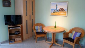 Een kamer met een televisie, een garderobe, rieten stoelen, een ronde tafel en een schilderij van een vuurtoren.