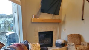 A living room with a fireplace, a television mounted above it, a sofa with patterned cushions, a wooden coffee table, and a dog on the floor. -    Enjoy the Summer Time in Views!  (Silverthorne)