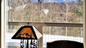 A room with a large window offering a view of a mountainous landscape. There is a table with a lamp and a chair in the foreground. -    Enjoy the Summer Time in Views!  (Silverthorne)