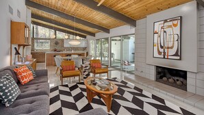 A modern living room with a fireplace, a dining area, and a kitchen visible through an open door. - Mid-Century Modern Gem - Pool Spa Fire Pit w/ Stunning Views in Vista Las Palmas (Palm Springs)