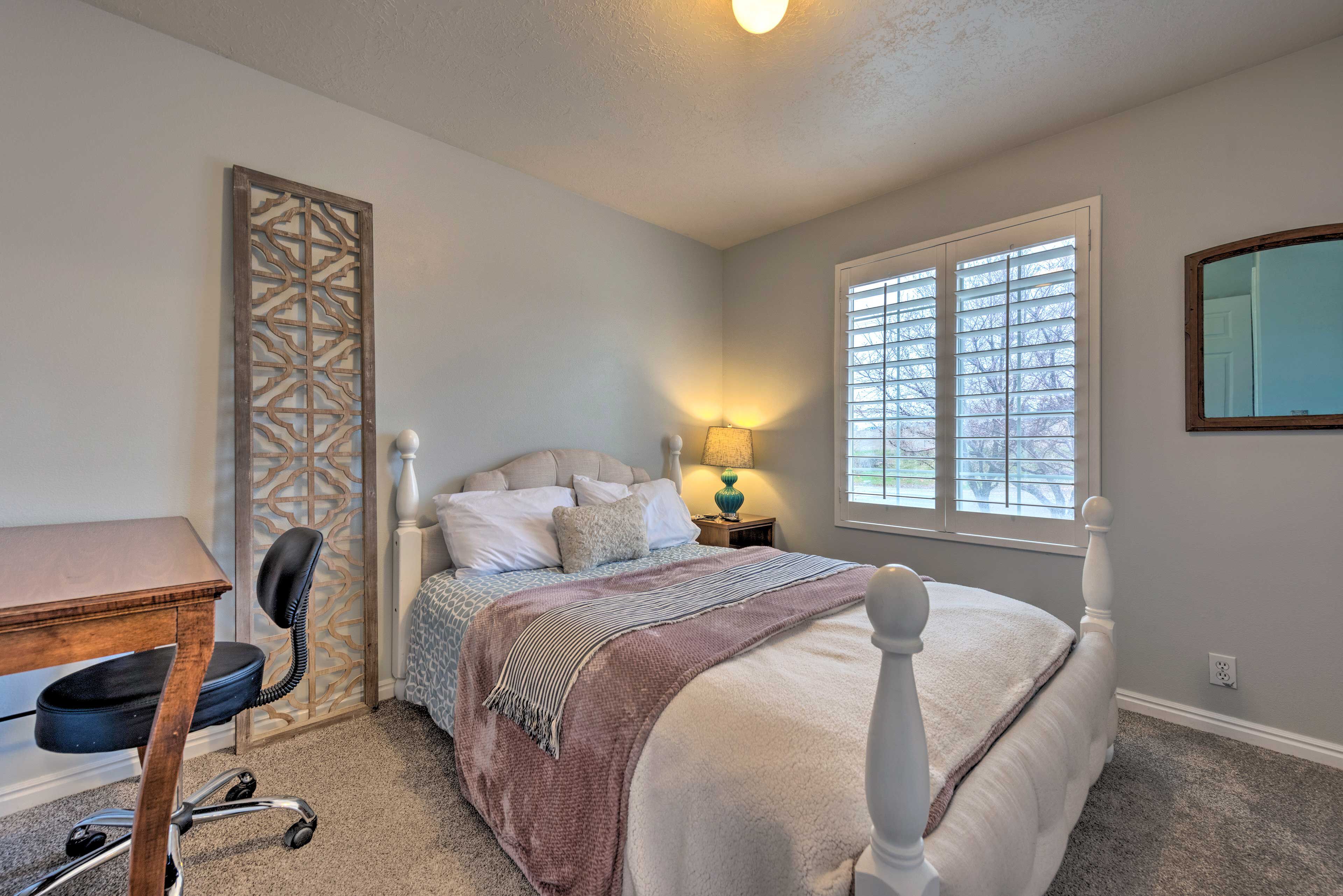 A bedroom with a bed, a desk, a chair, a window with blinds, and a decorative wall panel.