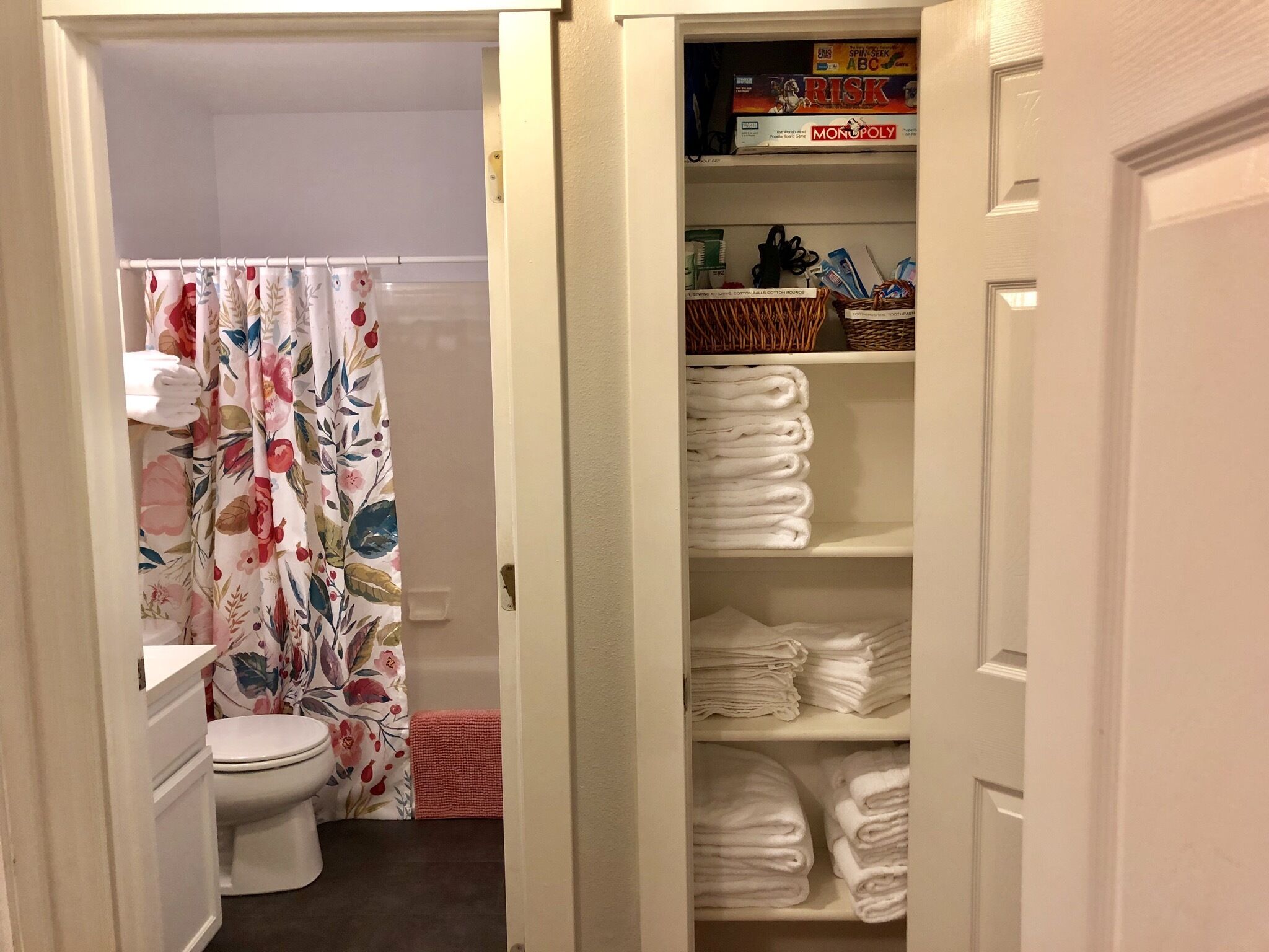 A bathroom with a toilet, a shower curtain with floral patterns, and a storage area with neatly stacked towels.
