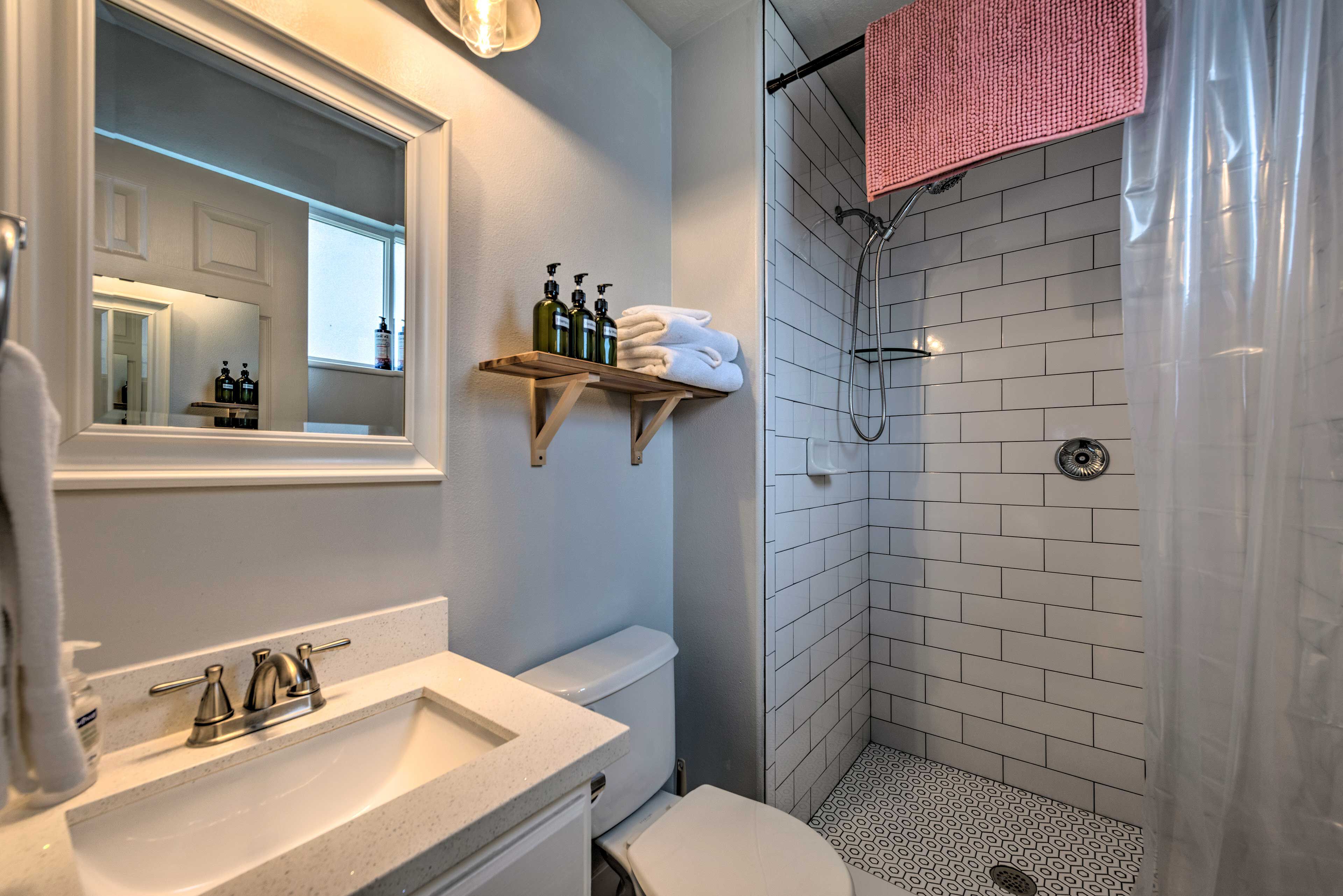 Un baño con lavamanos blanco, espejo y retrete. Hay una ducha con una toalla rosa colgada en la pared.
