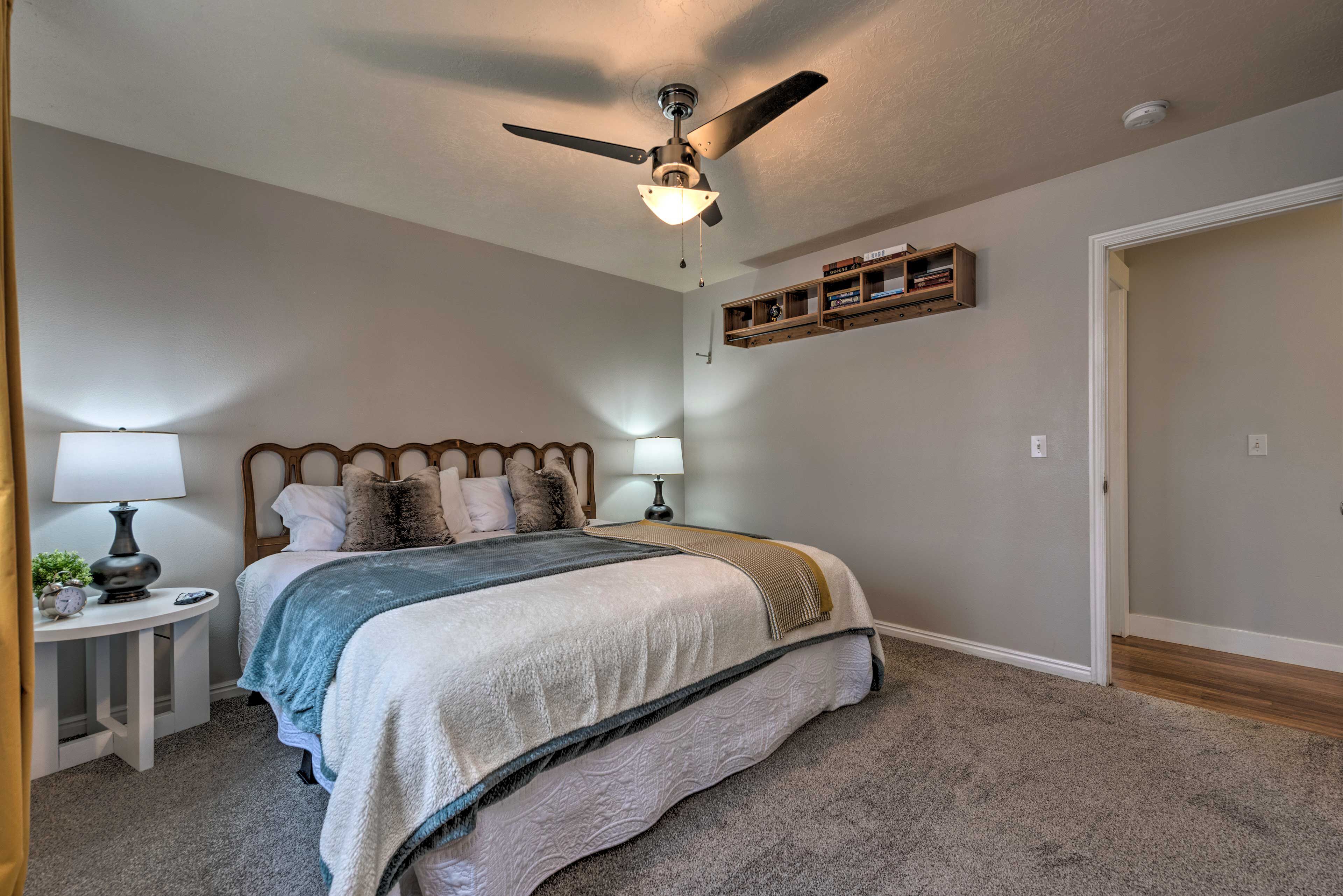 A bedroom with a bed, two bedside lamps, a ceiling fan, a nightstand, and a shelf with books.