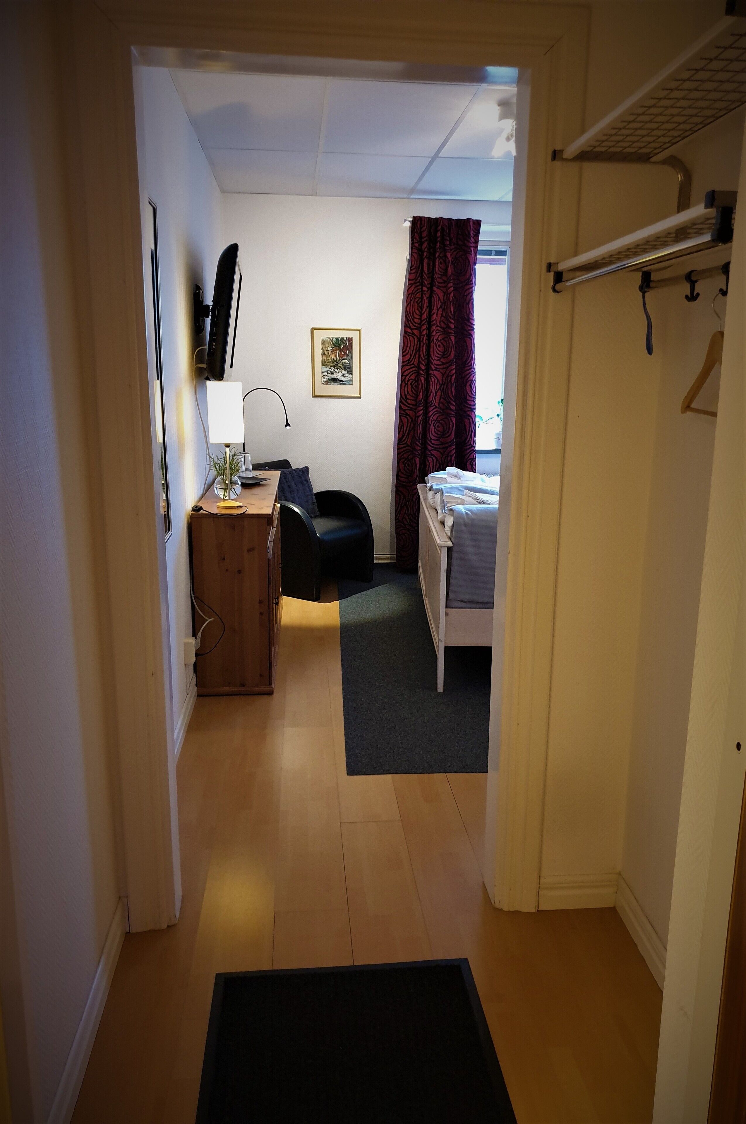 A narrow hallway leading to a bedroom with a bed, a desk, a chair, and a window with curtains.