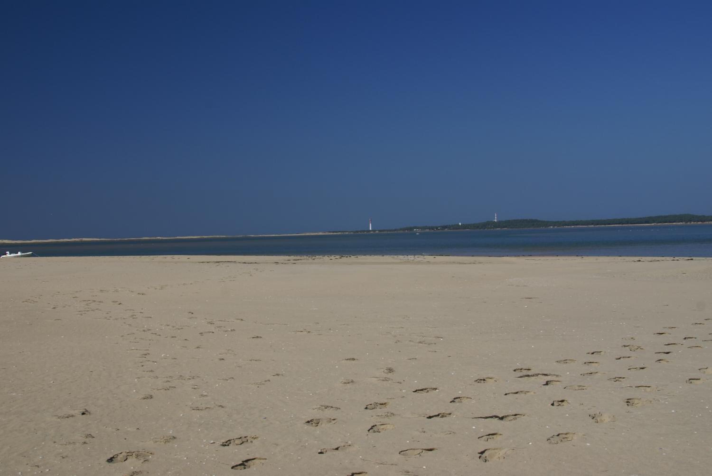 A sandy beach with footprints leading to the water, a distant lighthouse, and a small boat.