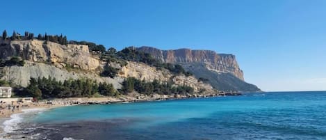 A coastal landscape with a rocky beach, clear blue sea, and a cliff with sparse vegetation.