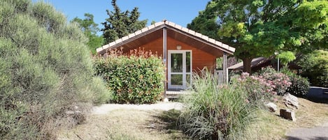 A small wooden cabin with a sloped roof, surrounded by dense shrubbery and trees.