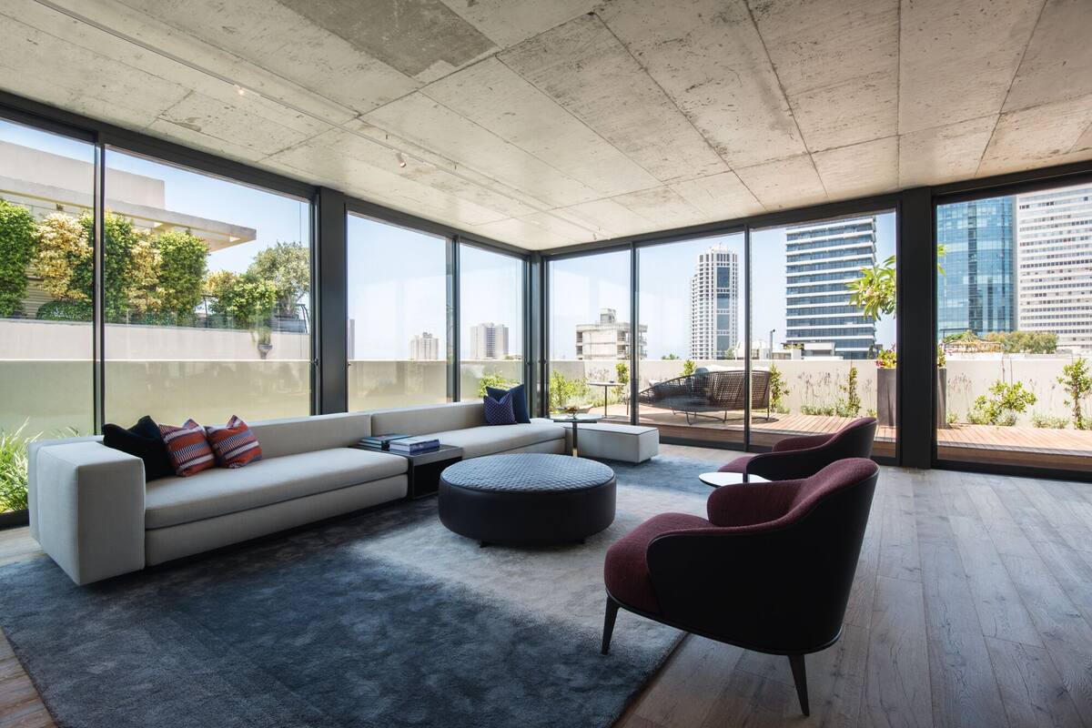 A modern living room with a sofa, ottoman, and chairs, large windows offering city views, and a concrete ceiling.