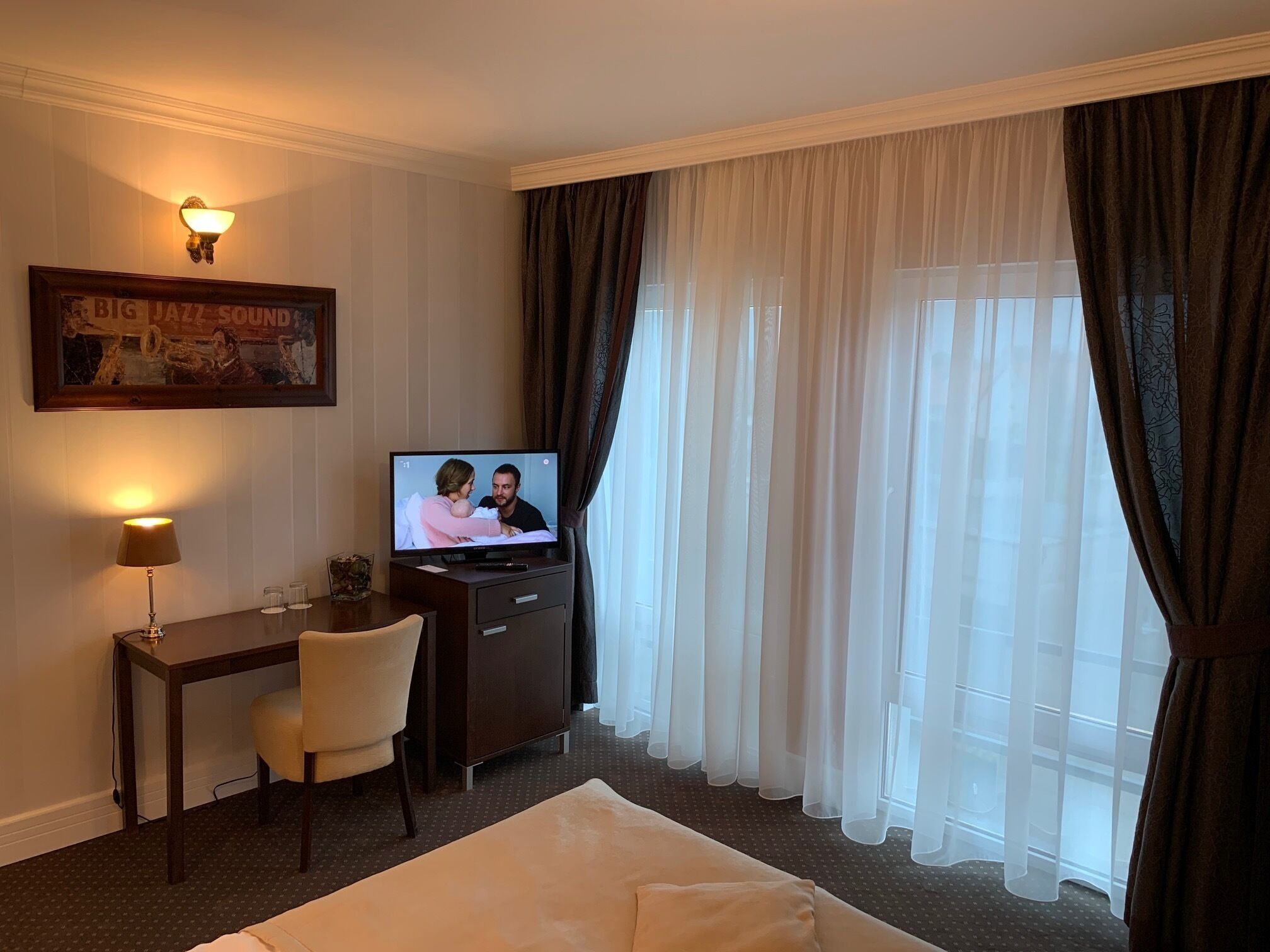 Een hotelkamer met een bed, een bureau met stoel, een televisietoestel en een groot raam met gordijnen.