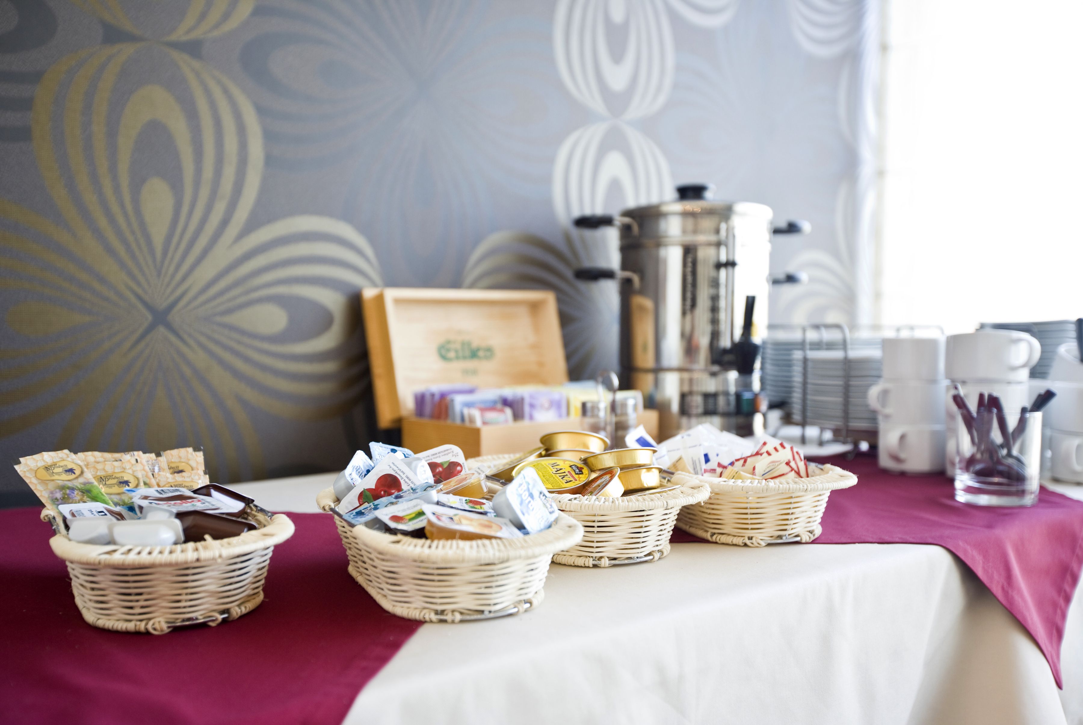 A breakfast buffet with wicker baskets filled with various food items, a coffee maker, and a framed menu on a table with a red tablecloth.