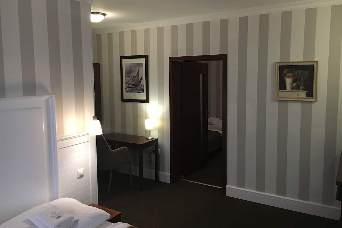 A hotel room with a bed, a desk with a chair, a framed picture on the wall, and a door leading to another room.