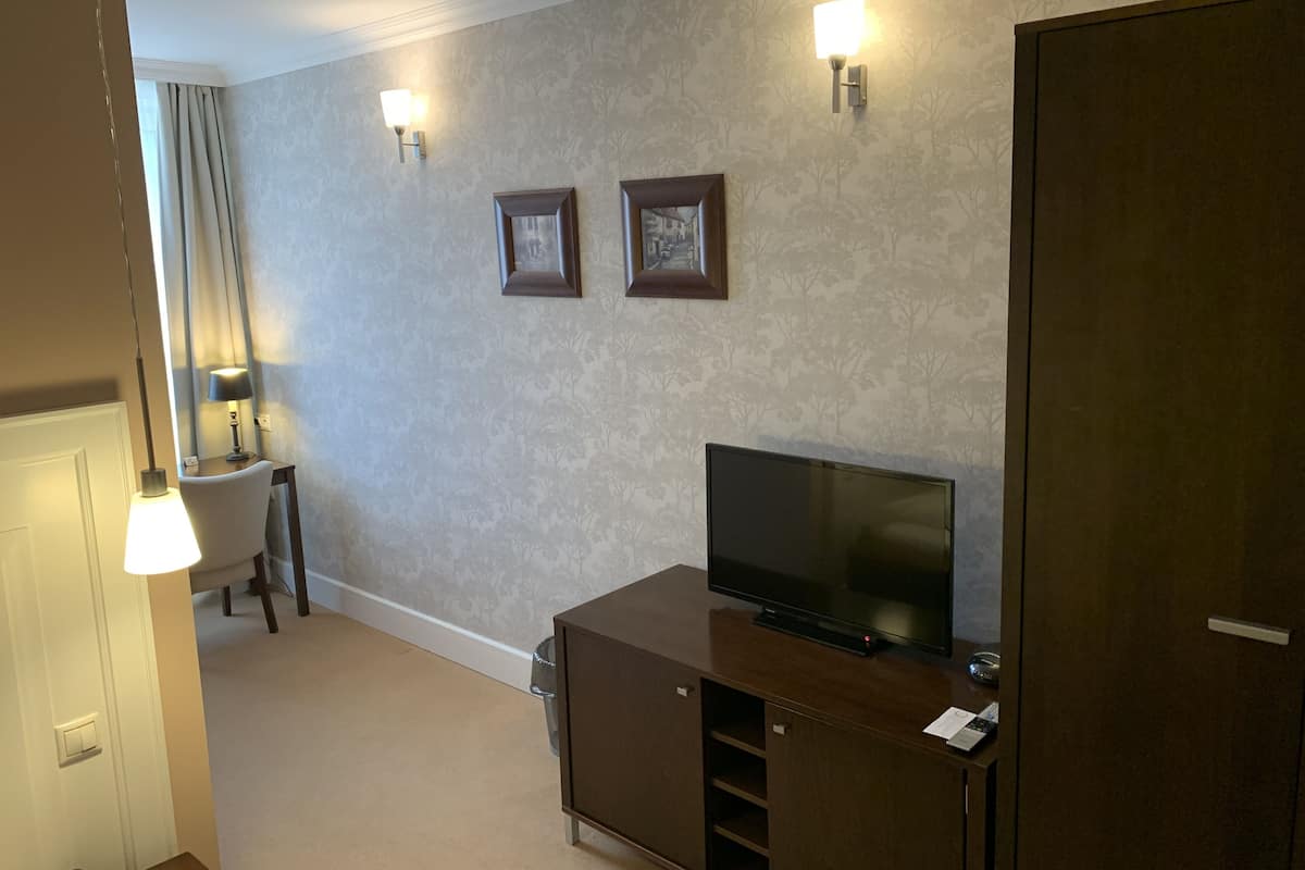 A hotel room with a flat-screen TV on a wooden cabinet, two wall-mounted picture frames, a lamp on a side table, and a door.