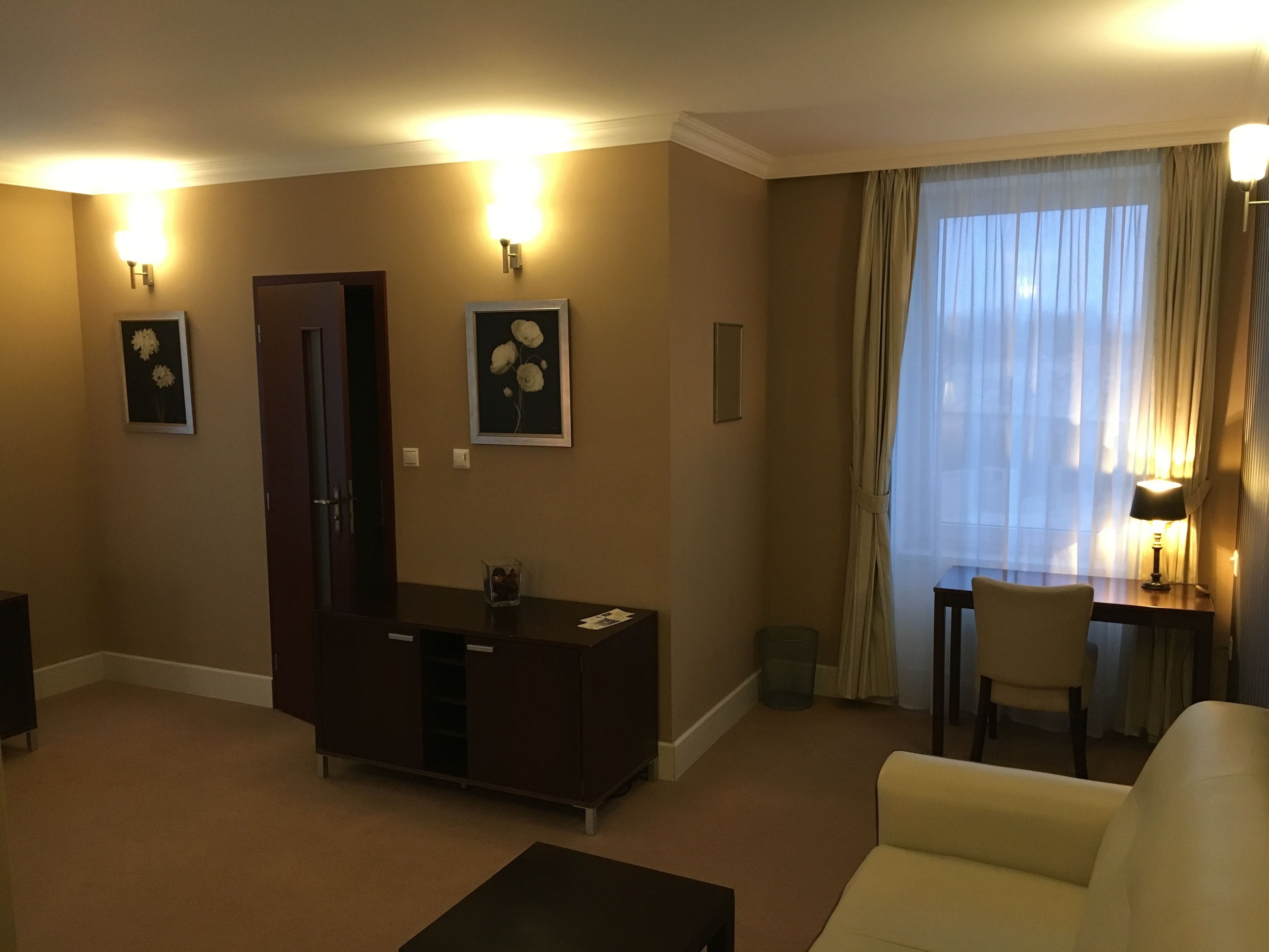 A hotel room with a sofa, a desk with a chair, a cabinet, and a large window with curtains.