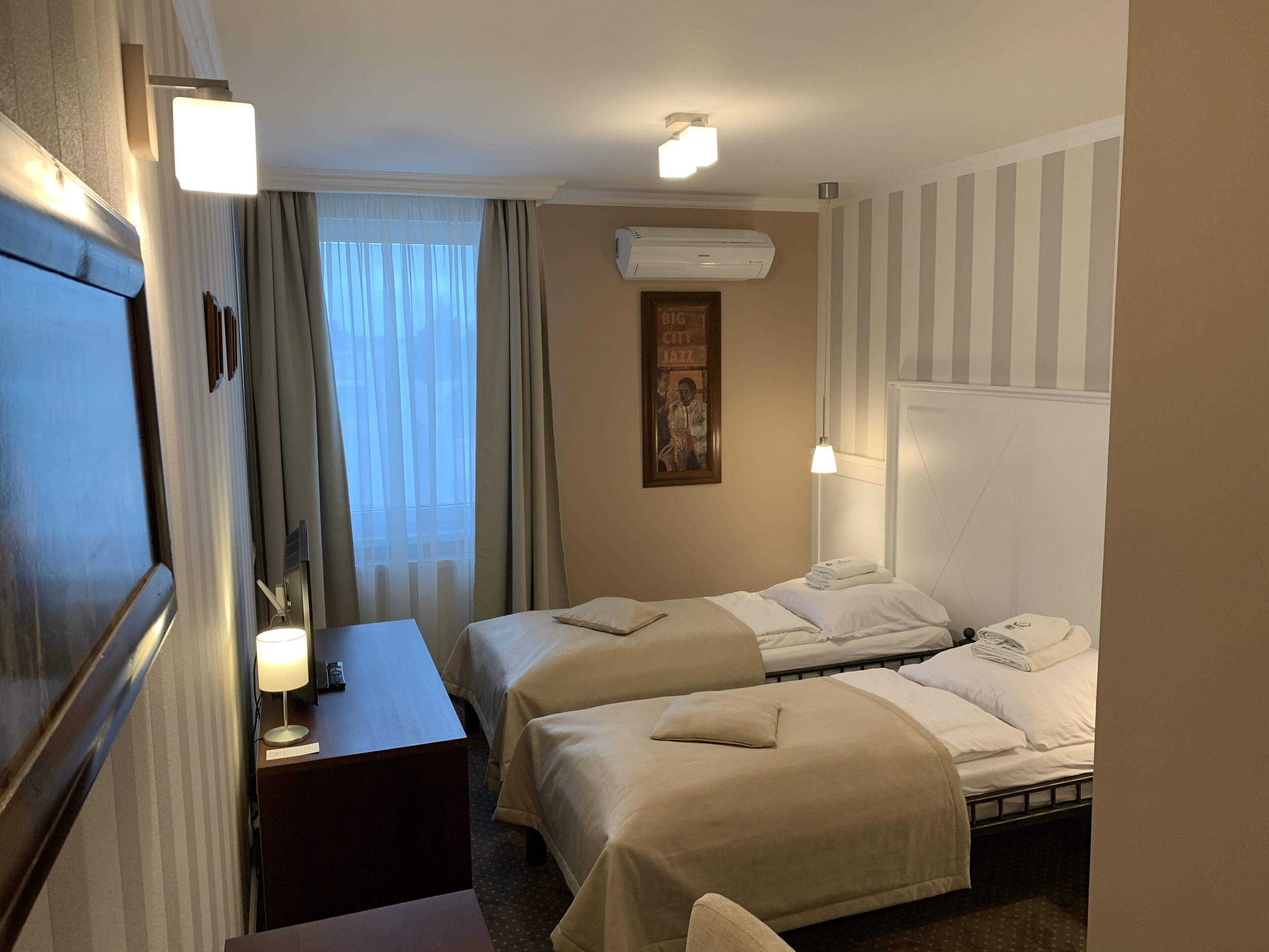 A hotel room with two beds, a desk, a lamp, an air conditioning unit, and a painting on the wall.