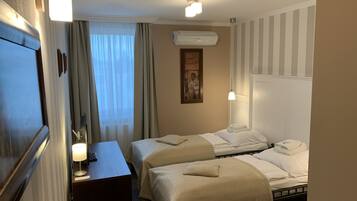 A hotel room with two beds, a desk, a lamp, an air conditioning unit, and a painting on the wall.