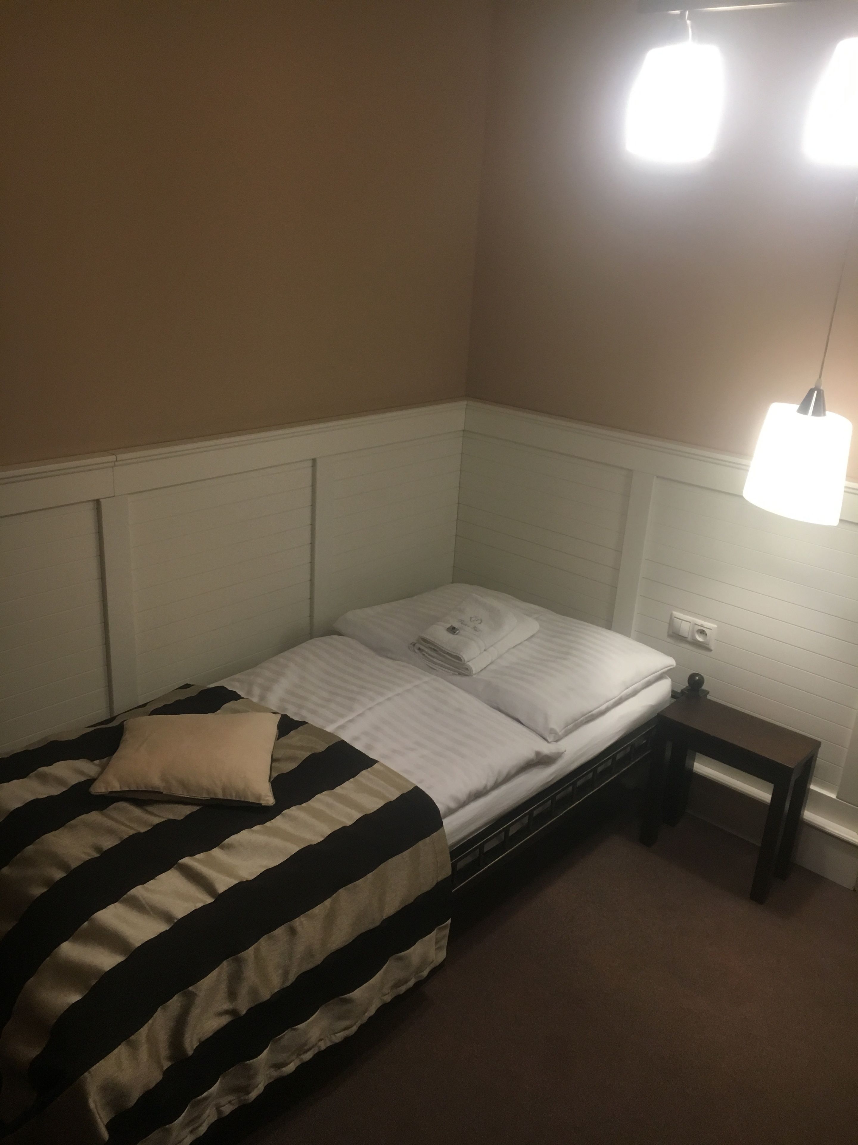 A single bed with a striped bedspread, a bedside table with a lamp, and a wall-mounted light fixture.
