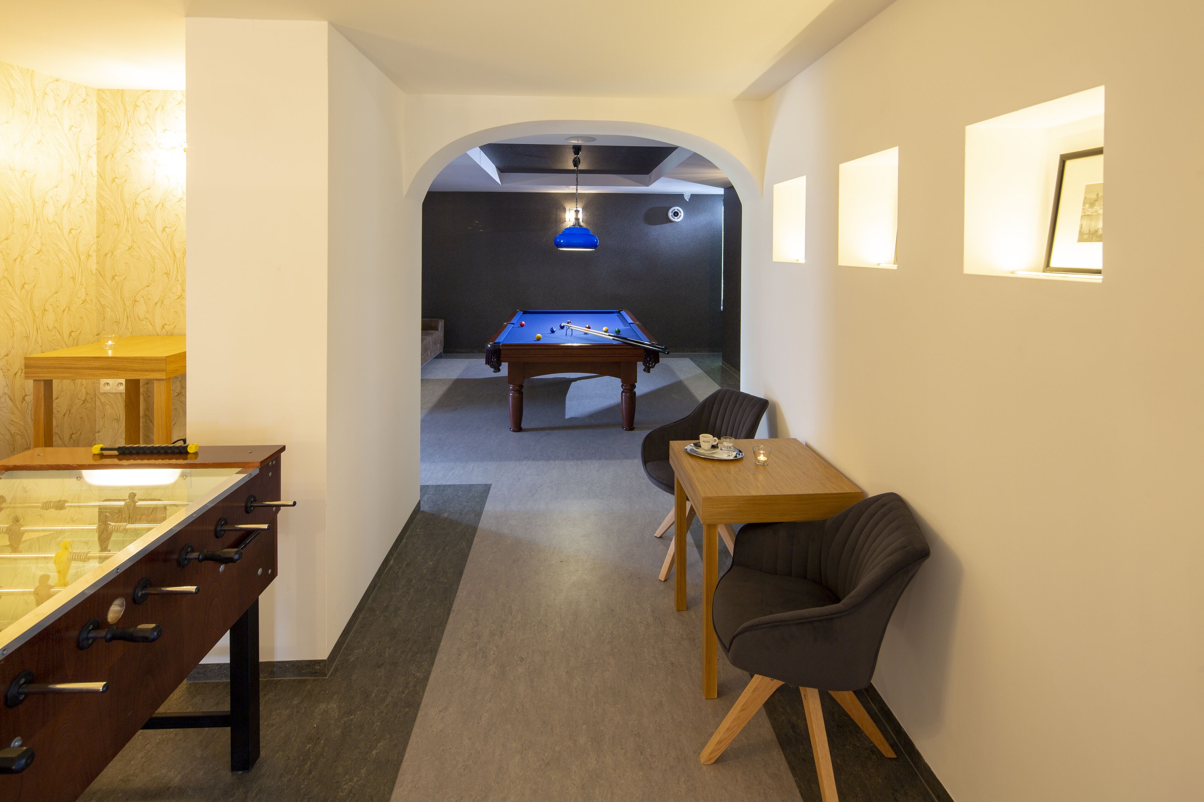 A room with a foosball table, a pool table, and a billiards table. There is a table with a plate and a cup, and a chair.