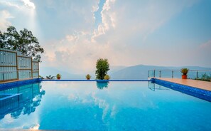 A clear blue pool with a wooden deck and potted plants, surrounded by a metal fence and trees, with a mountainous backdrop. - Cloud Mist Resort and Spa (Mahabaleshwar)