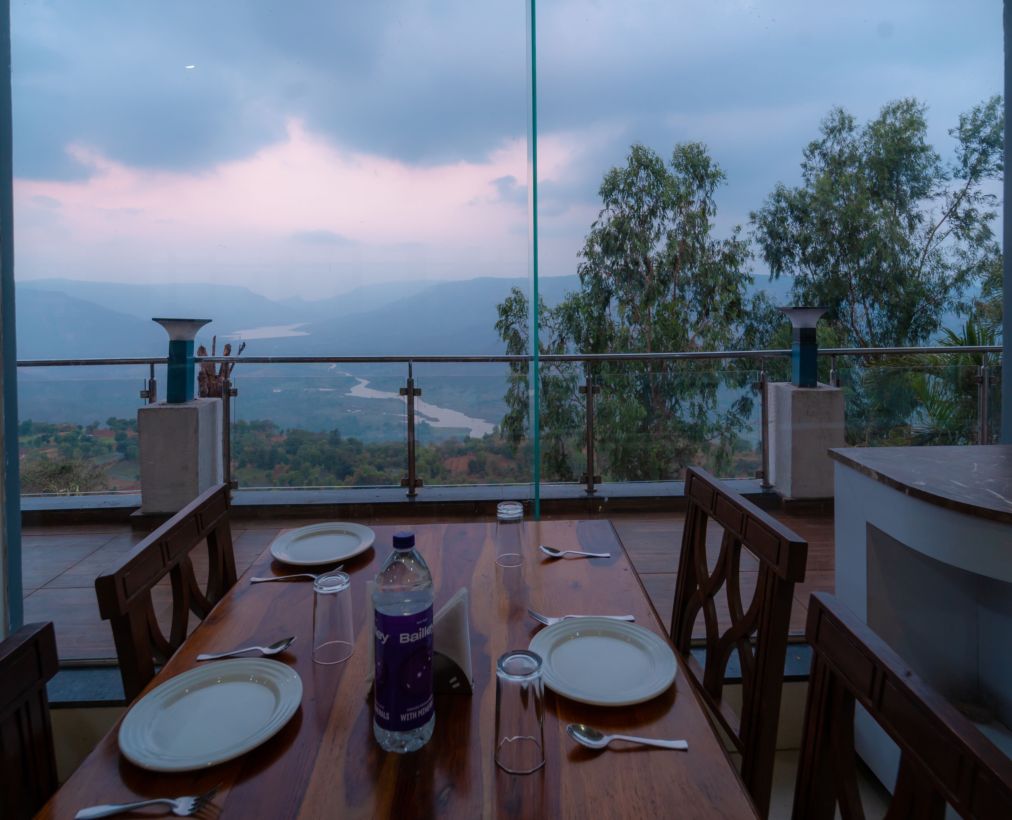 Une table à manger dressée pour deux personnes, avec une bouteille d’eau, des assiettes et des couverts. La table est placée sur un balcon d’où l’on peut admirer une rivière et des montagnes.