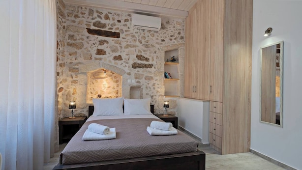 A bedroom with a stone wall, a wooden wardrobe, a bedside table with a lamp, and a bed with towels. - Melina Apartment in the heart of Rethymno Old Town (Rethymno)