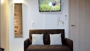 A modern living room with a brown sofa, two white cushions, a mounted flat-screen TV, and a stone-textured window. - Melina Apartment in the heart of Rethymno Old Town (Rethymno)