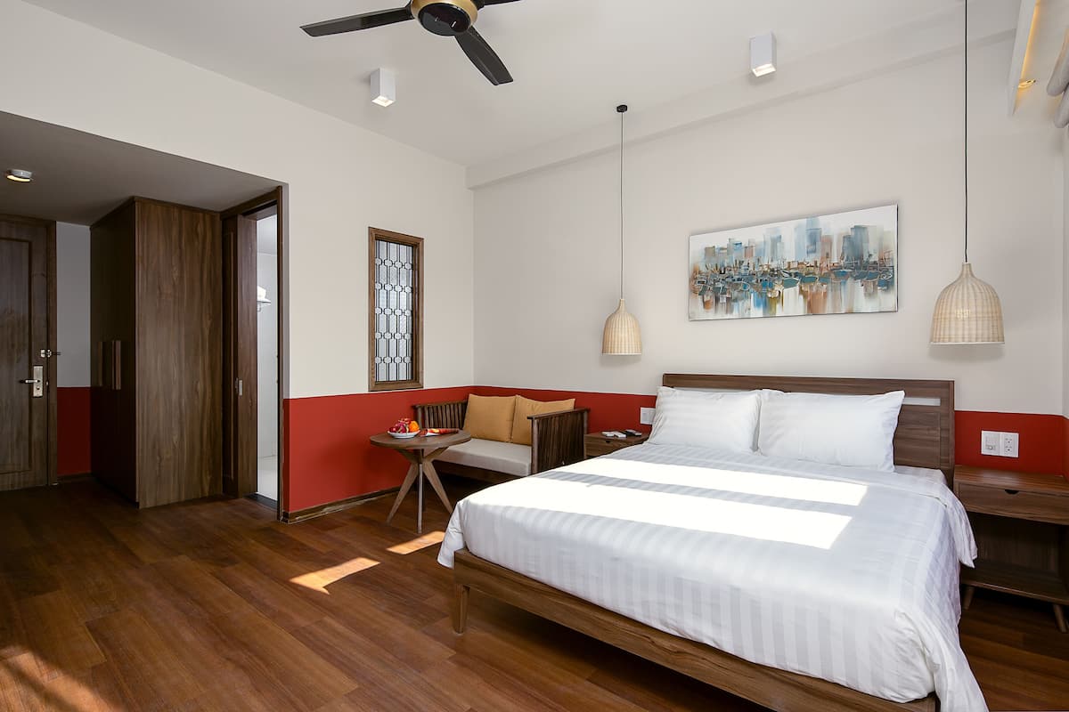 A modern hotel room with a large bed, a wooden floor, a red accent wall, a cityscape painting, and a ceiling fan.