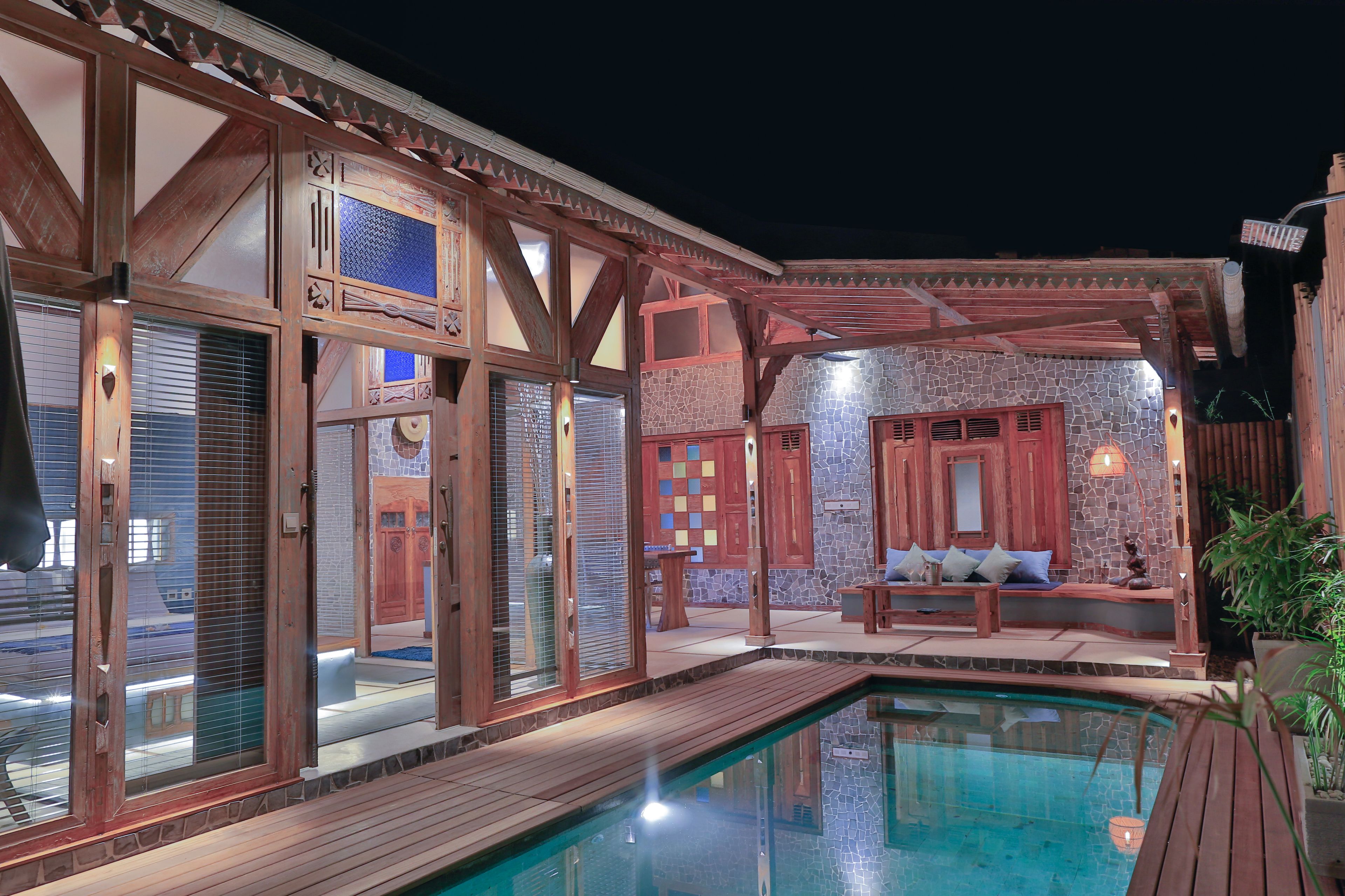 A pool area with wooden deck, stone walls, and a seating area with cushions.