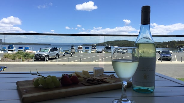 Una mesa con una botella de vino blanco, una copa, y una tabla de madera con uvas y queso. Se ve un puerto deportivo con barcos y coches al fondo.
