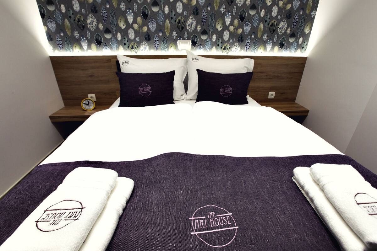 Una cama bien tendida con un cabecero de diseño floral, dos almohadas con un logotipo y una mesita de noche con un reloj.