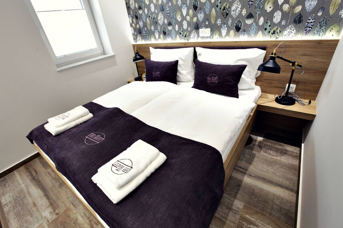Una cama con ropa de cama blanca y morada, un cabecero de madera y una mesita de noche con una lámpara.