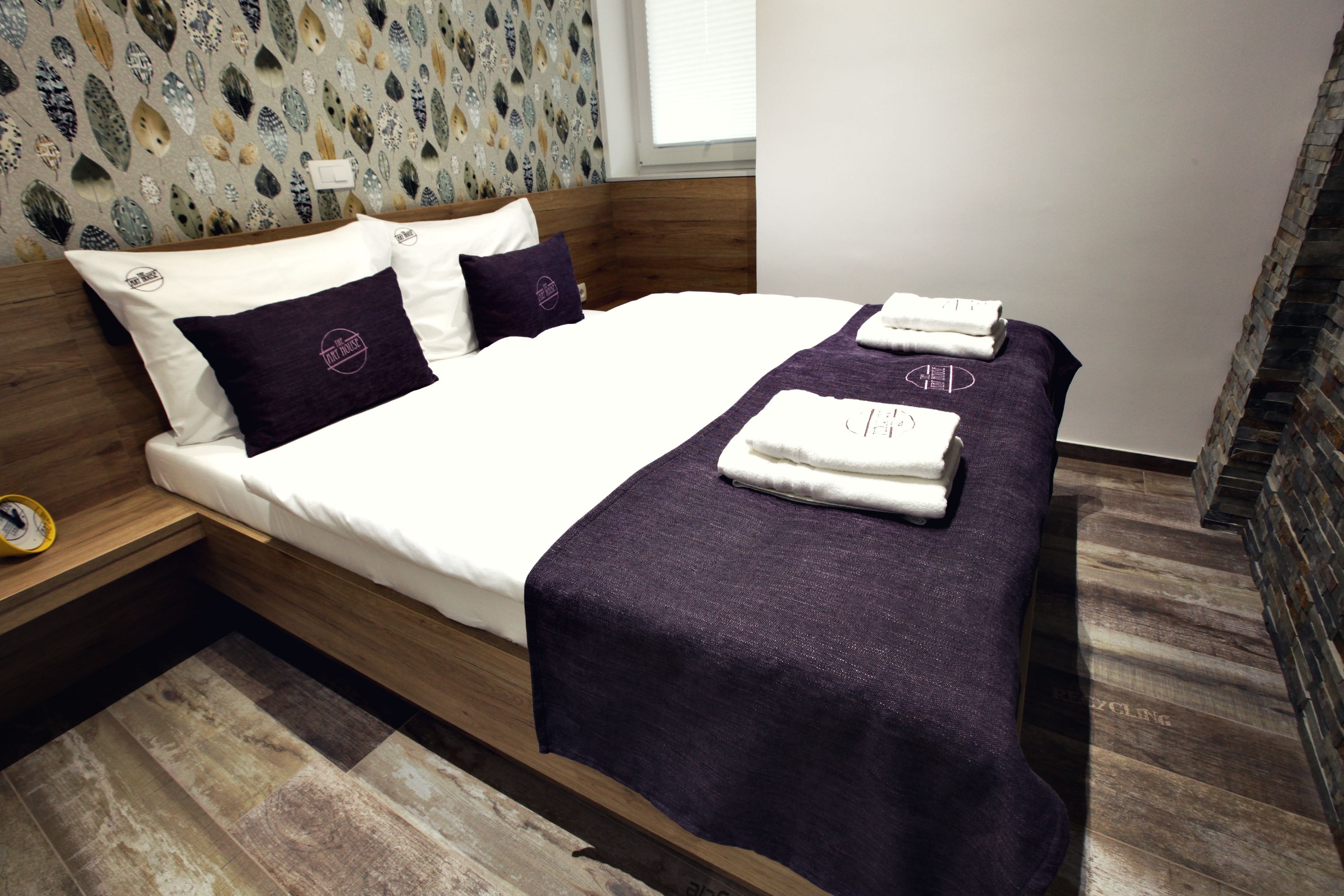 Una cama bien hecha con una manta y almohadas moradas, un cabecero de madera y un papel tapiz estampado.