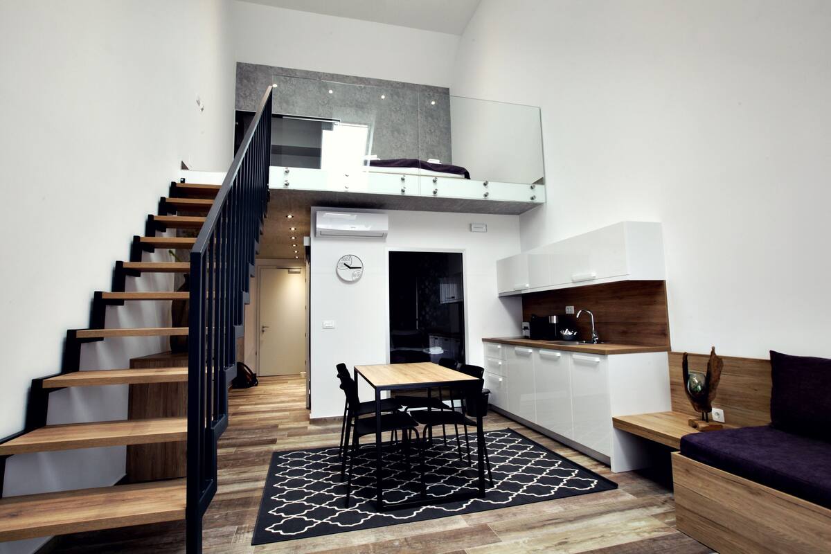 Un apartamento moderno con una escalera de madera, cocina con armarios blancos, zona de comedor y un dormitorio estilo altillo con cama y ventana grande.