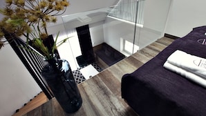 Una habitación de hotel moderna con una escalera de cristal, una cama con colcha morada y un jarrón con flores secas sobre un piso de madera.