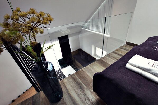 A modern hotel room with a glass staircase, a bed with a purple bedspread, and a vase with dried flowers on a wooden floor.