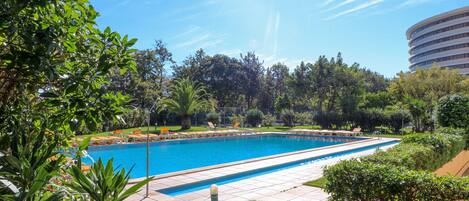 A swimming pool with a tiled deck, surrounded by greenery and a building in the background.