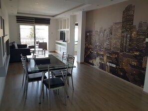 A modern dining area with a glass table and metal chairs, a kitchen with a refrigerator and TV, and a cityscape mural on the wall.